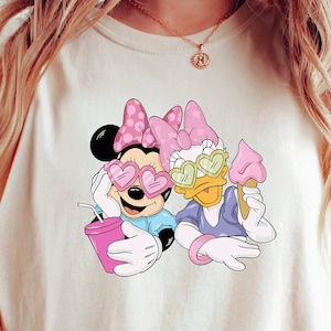 May include: A white oversized t-shirt with a graphic print of Mickey Mouse and Daisy Duck wearing heart-shaped sunglasses. The graphic also includes a pink drink and an ice cream cone.