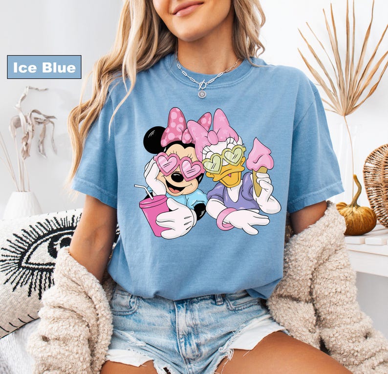 May include: Ice blue t-shirt featuring a graphic of Minnie Mouse and Daisy Duck wearing sunglasses and holding ice cream and a drink. The characters are in a relaxed pose, with a pink and purple color scheme.