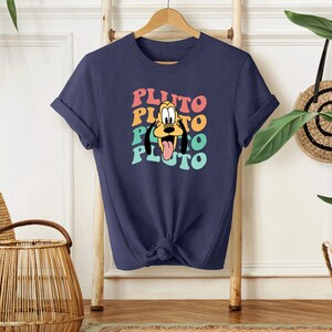 May include: A navy blue t-shirt with a retro-style graphic of Pluto, the Disney dog. The graphic features Pluto's face with a rainbow color scheme and the word "Pluto" repeated four times.
