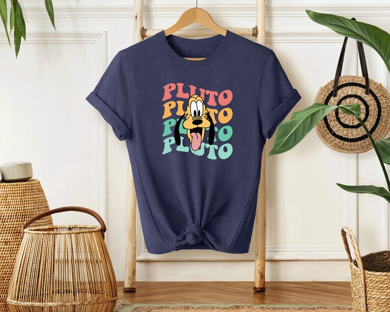 May include: A navy blue t-shirt with a retro-style graphic of Pluto, the Disney dog. The graphic features Pluto's face with a rainbow color scheme and the word "Pluto" repeated four times.