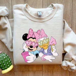May include: Beige crewneck sweatshirt featuring Minnie Mouse and Daisy Duck wearing heart-shaped sunglasses. Minnie holds a pink drink, and Daisy holds an ice cream cone. The graphic is in shades of pink, blue, and white.
