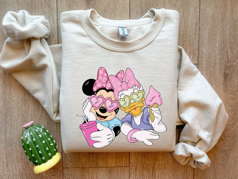 May include: Beige crewneck sweatshirt featuring Minnie Mouse and Daisy Duck wearing heart-shaped sunglasses. Minnie holds a pink drink, and Daisy holds an ice cream cone. The graphic is in shades of pink, blue, and white.
