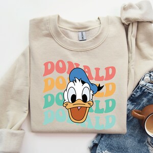 May include: A beige sweatshirt with a retro-style graphic of Donald Duck's face and the word "DONALD" repeated in colorful, layered text.