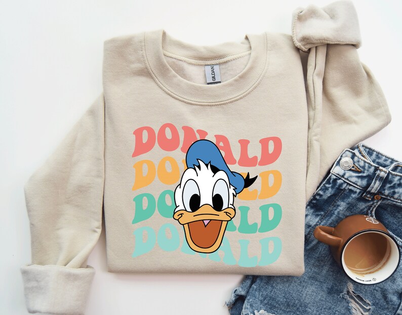 May include: A beige sweatshirt with a retro-style graphic of Donald Duck's face and the word "DONALD" repeated in colorful, layered text.
