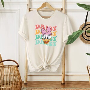 May include: A white t-shirt with a graphic of Daisy Duck's face and the word "Daisy" repeated four times in a rainbow color scheme.