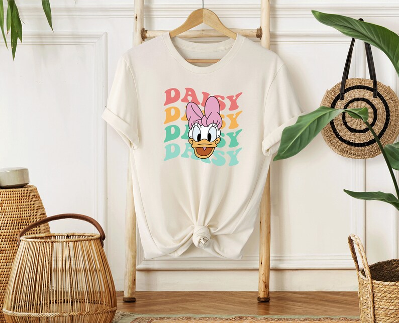 May include: A white t-shirt with a graphic of Daisy Duck's face and the word "Daisy" repeated four times in a rainbow color scheme.