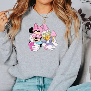 May include: A light gray sweatshirt featuring a cartoon graphic of Minnie Mouse and Daisy Duck wearing heart-shaped sunglasses. Minnie holds a pink drink, and Daisy holds an ice cream cone. The characters are in a relaxed pose.