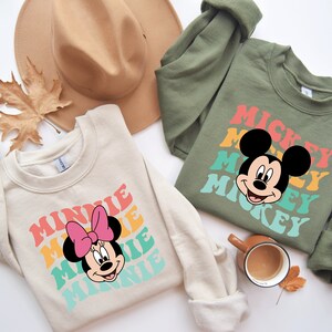 May include: Two cream colored sweatshirts with retro-style graphics of Mickey Mouse and Minnie Mouse. The Mickey Mouse sweatshirt is olive green and the Minnie Mouse sweatshirt is cream colored. The text on the sweatshirts reads "Mickey" and "Minnie" in a rainbow color scheme.