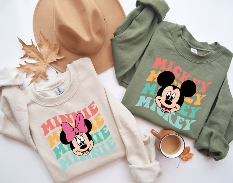 May include: Two cream colored sweatshirts with retro-style graphics of Mickey Mouse and Minnie Mouse. The Mickey Mouse sweatshirt is olive green and the Minnie Mouse sweatshirt is cream colored. The text on the sweatshirts reads "Mickey" and "Minnie" in a rainbow color scheme.