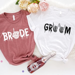 May include: Two t-shirts with a dripping font design. The pink shirt says "BRIDE" with a skeleton hand giving the middle finger. The white shirt says "GROOM" with two skeleton hands making the rock and roll sign.