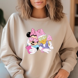 May include: Beige sweatshirt featuring a graphic of Minnie Mouse and Daisy Duck. Minnie Mouse is wearing heart-shaped sunglasses and holding a pink drink. Daisy Duck is holding an ice cream cone. Both characters are wearing pink bows.