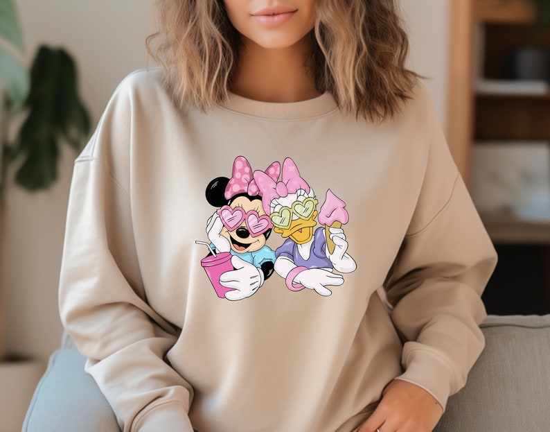 May include: Beige sweatshirt featuring a graphic of Minnie Mouse and Daisy Duck. Minnie Mouse is wearing heart-shaped sunglasses and holding a pink drink. Daisy Duck is holding an ice cream cone. Both characters are wearing pink bows.