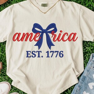 May include: A cream-colored short-sleeve t-shirt featuring the red text "ame" and "rica" separated by a large blue bow graphic. Below the bow, the blue text "EST. 1776" is printed. The shirt has a crew neckline and a visible brand tag at the collar.
