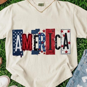 May include: Off-white t-shirt featuring the word "AMERICA" in a patchwork style. The letters are in red, white, and blue, with star details. A gold necklace is visible at the neckline. The shirt is on a grassy surface.