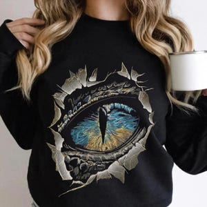 May include: A black long-sleeve sweatshirt featuring a graphic print of a large, detailed reptilian eye with a vertical pupil. The eye displays vibrant blue and gold iris tones, surrounded by a textured, cracked effect that appears to tear through the fabric.