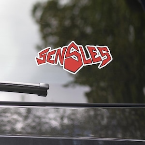 Könnte beinhalten: Ein rot-weißer Aufkleber mit dem Wort "SENSLES" in einer kräftigen, stilisierten Schriftart. Der Aufkleber befindet sich auf der Heckscheibe eines dunkelgrauen Autos, mit einem verschwommenen Hintergrund von Bäumen.