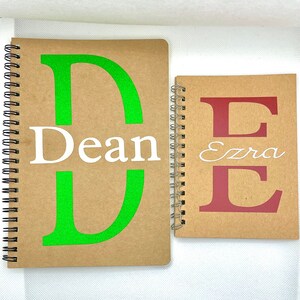 May include: Two brown spiral notebooks with personalised covers. The first notebook has a green letter "D" and the name "Dean" in white. The second notebook has a red letter "E" and the name "Ezra" in white.