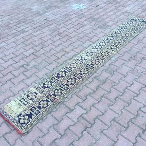 May include: A long, narrow rug with a geometric pattern in shades of beige, black, and red. The rug has a traditional design with repeating motifs.