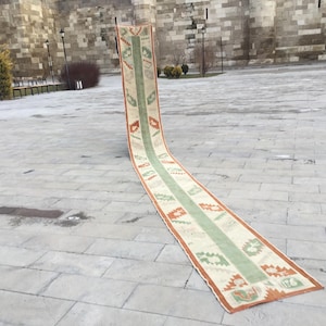 May include: A long, rectangular rug with a central green stripe and patterned borders in orange, green, and cream. The rug is displayed outdoors on a stone surface, showcasing its length and design.