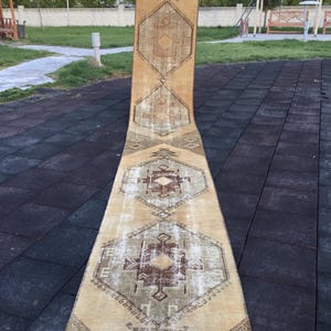 Może przedstawiać: Długi, beżowy dywanik z geometrycznym wzorem w odcieniach brązu, beżu i zieleni. Dywanik ma zużyty, vintage'owy wygląd.