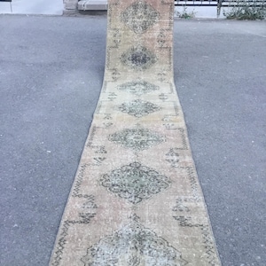May include: A long, rectangular rug with a faded, vintage appearance. The rug features a repeating geometric pattern in shades of green and gray on a beige background. The rug is displayed outdoors on a gray surface.