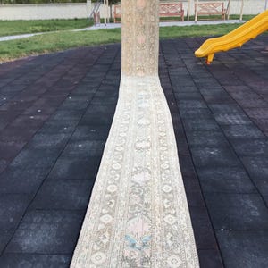 May include: A long, beige runner rug with a traditional floral pattern. The rug is laid out on a black rubber surface.