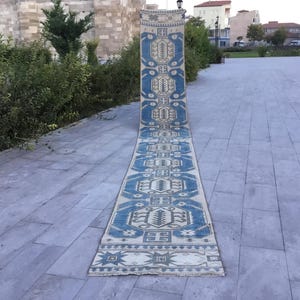 May include: A long, rectangular rug with a blue and cream geometric pattern. The rug is displayed outdoors on a stone surface, showcasing its intricate design and color palette. The rug's length is emphasized in the image.