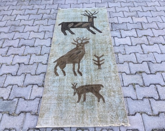 May include: A rectangular area rug featuring a faded light blue and tan background with three dark brown deer figures and a small tree motif. The rug has a distressed, weathered appearance with visible woven texture and short fringe on the ends.