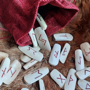 May include: A red drawstring bag spills out a collection of rune stones. The stones are off-white with red markings. The bag and stones rest on a bed of brown fur. The runes are a form of ancient writing.
