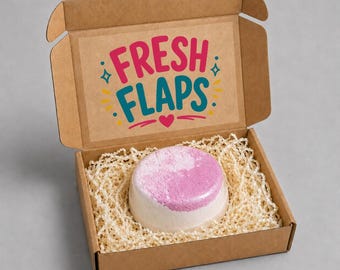 Könnte beinhalten: Eine runde, weiß-rosa Badebombe in einer braunen Pappschachtel, die mit Papierstreifen gefüllt ist. Auf der Innenseite des Deckels steht der Text FRESH FLAPS in Pink und Türkis, verziert mit kleinen gelben und türkisfarbenen grafischen Sternen.