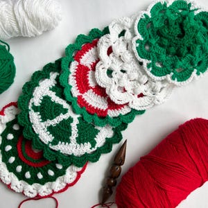 May include: A collection of handmade crochet coasters in red, white, and green. The coasters feature a scalloped edge and intricate designs. White and green yarn balls are visible in the background, along with a wooden crochet hook and a red yarn ball.