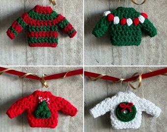 Puede incluir: Cuatro adornos navideños de crochet con forma de suéteres, cada uno con un diseño diferente. Los suéteres son rojos y verdes con detalles blancos. Los adornos están colgados de una cinta roja.