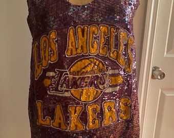May include: A purple sequined vest top featuring the words "LOS ANGELES LAKERS" in gold lettering and a gold basketball graphic. The top is displayed on a mannequin.