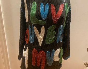 Puede incluir: Una blusa negra de lentejuelas con las palabras "I love you much" escritas en lentejuelas de colores.