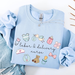 May include: Light blue sweatshirt with a graphic design of baby-related items and the text "labor & delivery nurse".