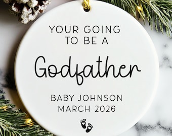 May include: A white ceramic ornament with the text "YOUR GOING TO BE A Godfather" in black. Below the text is "BABY JOHNSON MARCH 2026" and two small footprints. The ornament is hanging with a gold string.