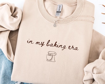 May include: A beige crewneck sweatshirt with the phrase "in my baking era" and a mixer graphic in brown. A silver necklace with a flower pendant is on the sweatshirt. The sweatshirt is folded on top of a pair of blue jeans and a beige sweater.
