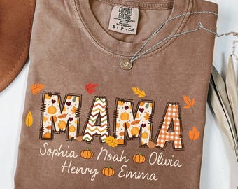May include: Brown Comfort Colors t-shirt with the word "MAMA" in a fall-themed design, featuring pumpkins and leaves. The names Sophia, Noah, Olivia, Henry, and Emma are printed below. A silver necklace with a pendant is draped across the shirt.