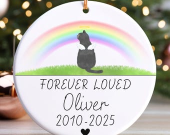 May include: A white ceramic ornament with a gold string for hanging. It features a black cat with angel wings and a halo, set against a rainbow and green grass. The text reads "FOREVER LOVED Oliver 2010-2025" with a heart.