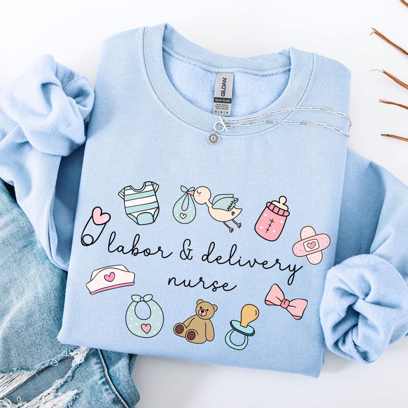 May include: Light blue sweatshirt with a graphic design of baby-related items and the text "labor & delivery nurse".