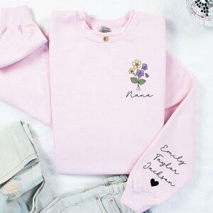 May include: Pink sweatshirt with a floral design and the word "Nana" on the front. The sleeve has the names "Emily, Taylor, Jackson" and a black heart.