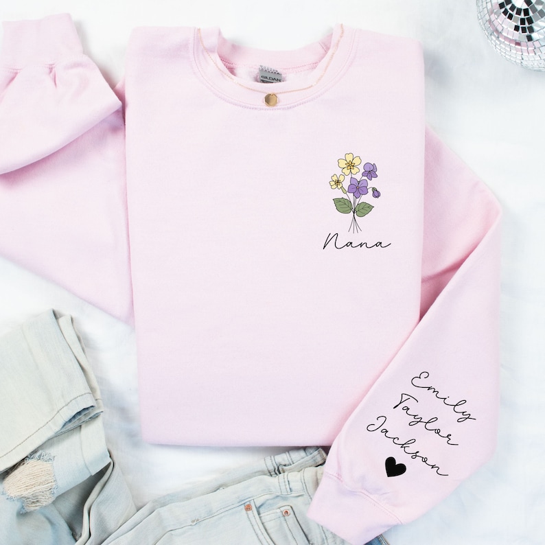 May include: Pink sweatshirt with a floral design and the word "Nana" on the front. The sleeve has the names "Emily, Taylor, Jackson" and a black heart.