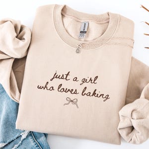 May include: A beige sweatshirt with the text "just a girl who loves baking" in brown script. A silver necklace with a flower pendant is draped across the top. The sweatshirt is folded on a white surface.