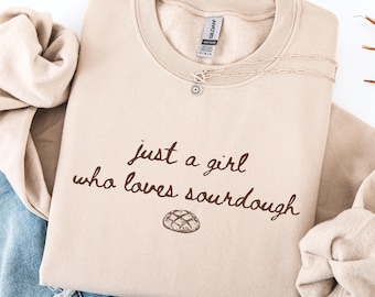 May include: A beige crewneck sweatshirt with the text "just a girl who loves sourdough" in brown script. A small illustration of a loaf of bread is below the text. A silver necklace with a flower pendant rests on the sweatshirt.