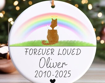 May include: A white ceramic ornament with a gold string for hanging. The ornament features a brown cat with angel wings and a halo, set against a rainbow. Text reads "FOREVER LOVED Oliver 2010-2025" with a black heart.