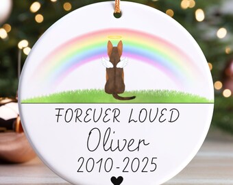 May include: White ceramic ornament with a gold string for hanging. The ornament features a brown and black dog with angel wings and a halo, sitting under a rainbow. Text reads "FOREVER LOVED Oliver 2010-2025" with a black heart.
