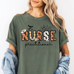 May include: Olive green t-shirt with a Halloween-themed design. The design features the word "NURSE" in orange and black, with a spider web, bats, and medical symbols. The word "practitioner" is written below.