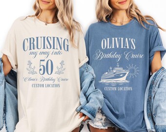 May include: Two t-shirts, one cream and one blue, with cruise-themed designs. The cream shirt says "Cruising my way into 50" with anchors. The blue shirt says "Olivia's Birthday Cruise" with a ship graphic. Both shirts have "Custom Location" text.