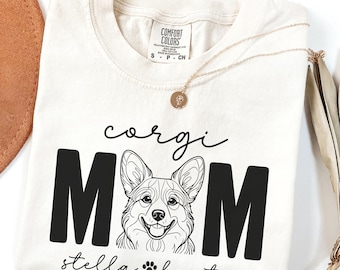 May include: Cream-colored t-shirt with the words "corgi MOM" in black block letters and a corgi illustration. The name "stella baxter" is below the illustration, with a paw print. A gold necklace is draped on the shirt.