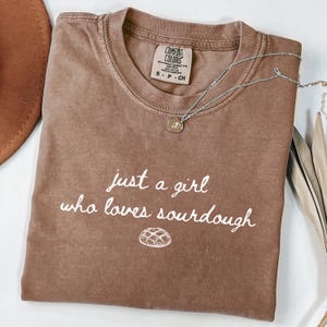 May include: A brown Comfort Colors t-shirt with the text "just a girl who loves sourdough" in white script. A small illustration of a loaf of bread is below the text. A silver necklace with a small pendant is on the shirt.